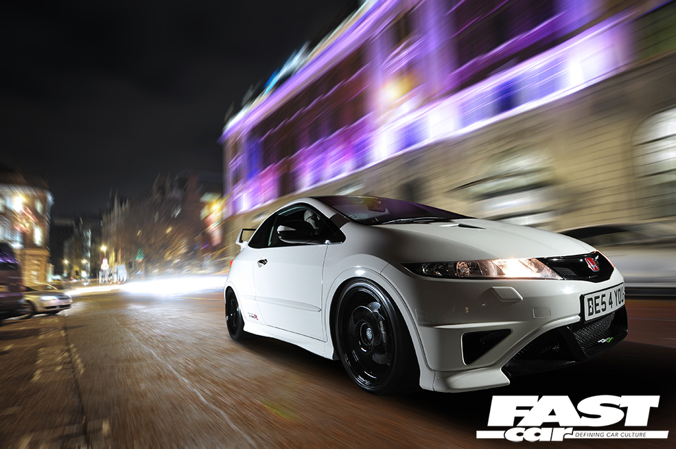 A close up front right shot of white Honda Civic FN2 TYPE-R TURBO 00 driving in city with blurred lights behind